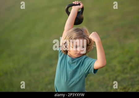 Child lifting the kettlebell in park outside. Cute child boy pumping up arm muscles with kettlebell. Fitness kids with dumbbells Stock Photo