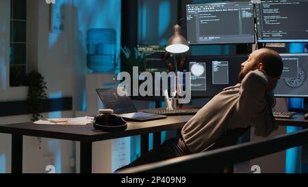Smiling coder feeling content about programming achievement, developing new user interface at night. Young adult celebrating IT success and being cheerful or pleased, laid back in office. Stock Photo