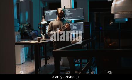 Young programmer playing air guitar in agency office, enjoying music and song late at night. Having fun fooling around, feeling cheerful and careless while he programs new code. Handheld shot. Stock Photo