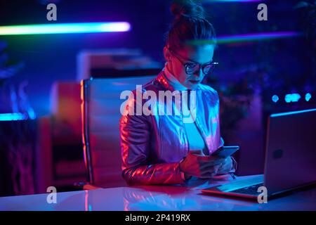 Neon metaverse futuristic concept. elegant woman in glasses with laptop using smartphone in modern office. Stock Photo