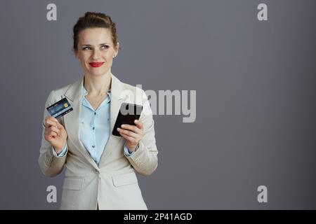 smiling elegant female employee in a light business suit with smartphone and credit card isolated on gray. Stock Photo
