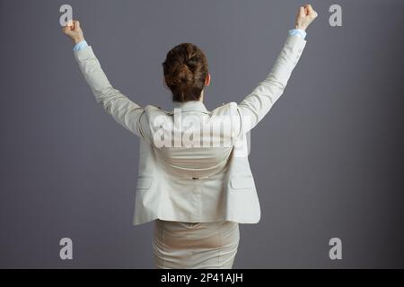 Seen from behind female worker in a light business suit with raised arms rejoicing isolated on gray. Stock Photo