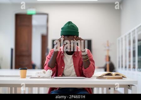 Tired Black student man sitting in library feeling unmotivated to study and trying to concentrate. Stock Photo