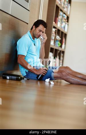 Hes inconsolable. a crying man drinking wine while sitting on the floor with some tissues. Stock Photo