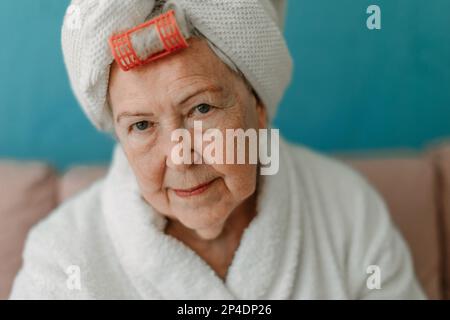 Happy senior woman with plastic curler sitting at sofa in bathrobes and looking at camera. Stock Photo