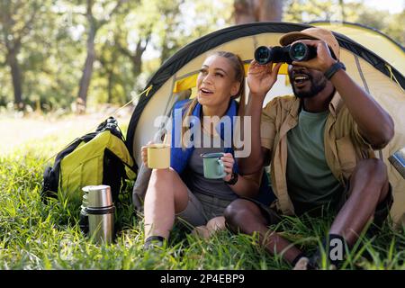 Happy, diverse couple in forest sitting in front of tent with coffee and using binoculars Stock Photo
