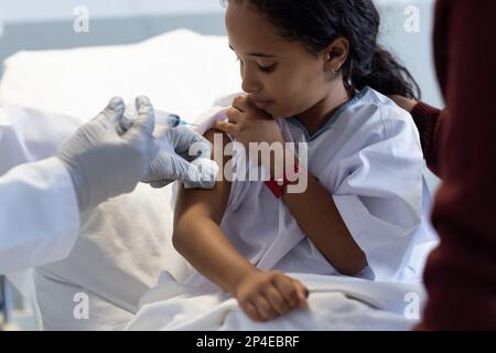 Diverse doctor vaccinating sick daughter with mother on hospital bed Stock Photo