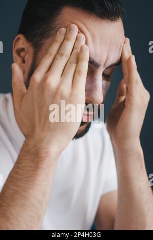 Close-up portrait of frustrated handsome young man touching his temles and keeping eyes closed while sitting on the couch at home. Exhausted, Illness Stock Photo