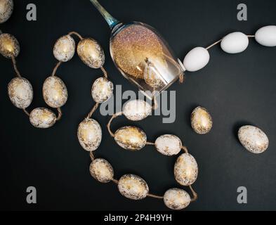 Creative , matt black background with white and gold easter eggs attached to the rope. Wine glass with golden sparkles. Flat lay, bird view. Stock Photo