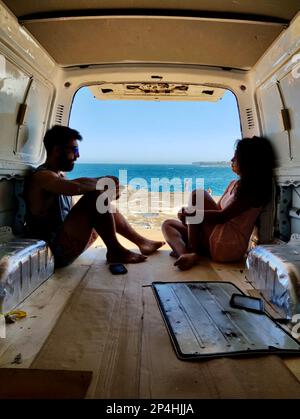 Relaxing couple seated inside the back of a van in front of the ocean. Stock Photo