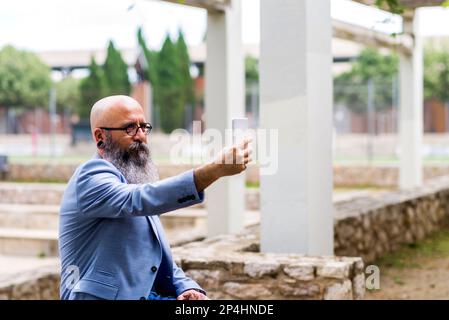 middle-aged, bald, bearded man sitting taking selfie in park Stock Photo