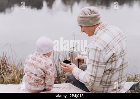 Dad and baby daughter are sitting by the river and drinking tea Stock Photo