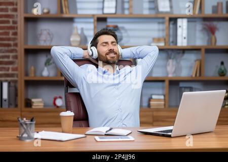 Mature businessman in blue shirt relaxing in office, man with headphones folded hands on title, listening to soothing music and audiobook podcasts, boss at workplace sitting using laptop. Stock Photo
