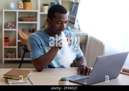 Young African American businessman smoking electronic cigarette while sitting by workplace in front of laptop and working over project Stock Photo