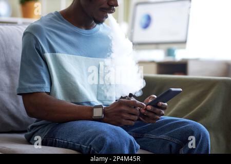 Cropped shot of African American guy with vape pen using mobile phone while sitting on couch in home office during break and relaxing Stock Photo
