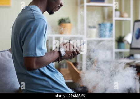 Side view of young black man in t-shirt standing in front of camera in living room and texting in smartphone while smoking e-cigarette Stock Photo