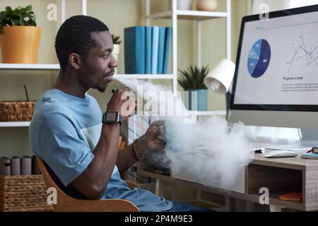 Side view of young businessman smoking e-cigarette and analyzing online graphic data on computer screen while sitting by workplace Stock Photo