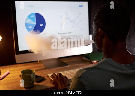 Young businessman smoking e-cigarette while sitting in front of computer with graphic data on screen and working late in the evening Stock Photo