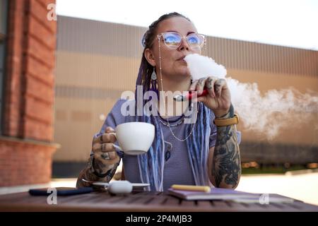 Young woman with tattoos on arms relaxing in outdoor cafe while having cup of coffee and blowing cloud of smoke out of her mouth Stock Photo