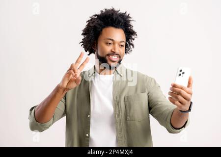 Handsome attractive 30s guy taking selfie by smartphone, showing peace symbol into webcam standing isolated on white, charismatic african man making video call, using mobile app for virtual connection Stock Photo