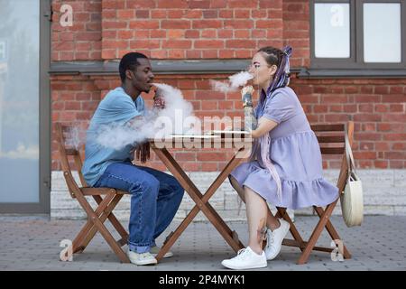 Side view of two young intercultural colleagues smoking electronic cigarettes while sitting by table and organizing work at meeting Stock Photo