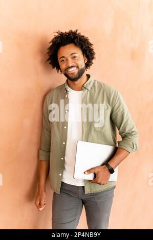 Vertical portrait of young african american guy standing isolated on wall holding laptop, looking camera, going on work, showing positive emotions. Technology and people concept Stock Photo