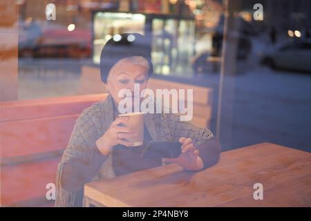 The hands of an elderly woman in a cafe, a pensioner drinking coffee and looking at the phone, talking on a mobile phone with friends and relatives. Front view. Stock Photo
