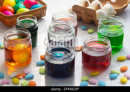 Dyeing Easter Eggs in Colorful Dye in Glass Jars with Multicolored Easter Candy on Marble Counter Stock Photo