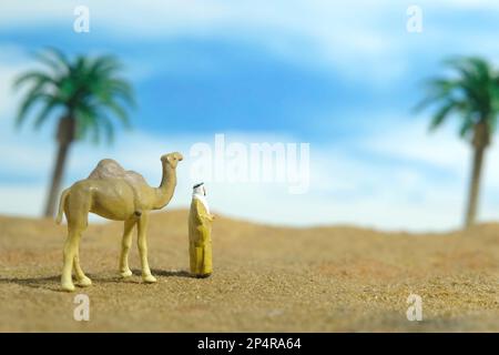 Miniature people toy figure photography. Middle eastern man wearing traditional clothes walking with camels in desert. Image photo Stock Photo