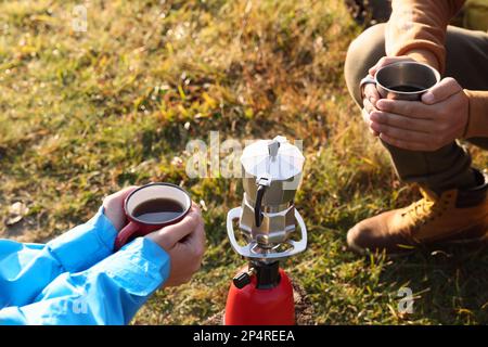 Tourists with mugs of coffee sitting around camping burner outdoors, closeup Stock Photo