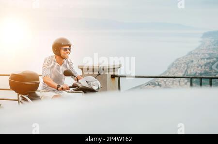 Cheerfully smiling man in helmet and sunglasses fast riding the moto scooter on the Sicilian old town streets on the Ionian Seaside coast. Happy Itali Stock Photo