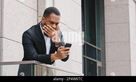 Frustrated ethnic African American bearded sad shocked man looking at smartphone screen feeling disappointed with received bad news message stress Stock Photo