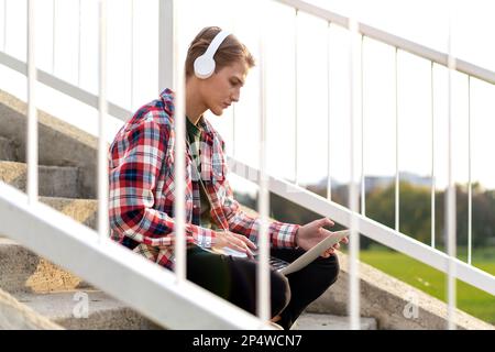 Woman student in headphones sitting on the steps in the city and using her laptop. Stock Photo