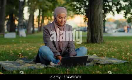 Muslim islamic ethnic student girl in hijab business woman freelancer worker uses laptop sitting on green grass in park working studying remotely in Stock Photo