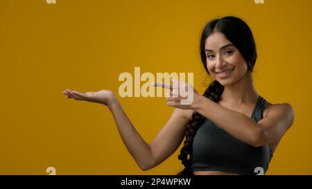 Portrait in studio Indian 20s woman posing on yellow background pointing to side at palm arm hand point aside recommend recommendation sign girl lady Stock Photo