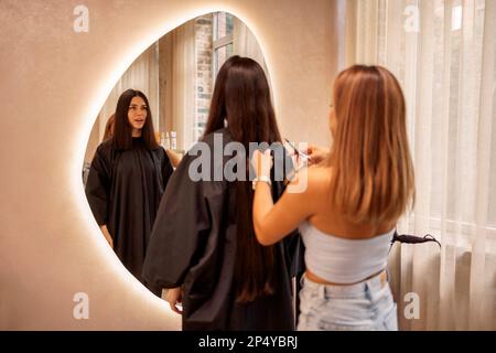 Blonde hairdresser cuts her female client hair in a beauty salon. Pretty brunette young woman looks into a huge mirror when her luxurious long hair is Stock Photo