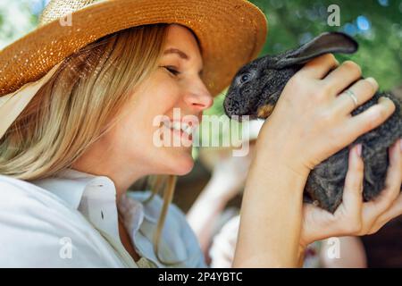 Close up of a young beautiful woman holding a small black rabbit. Caucasian pretty girl in a straw hat and a white shirt smiles and holds a cute bunny Stock Photo