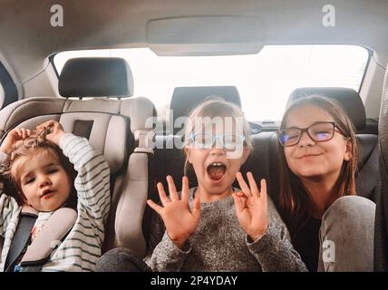 Three cute sisters of different ages are riding in the back seat of a car. Girls scream, indulge and laugh. Road trip with three children. Kids having Stock Photo