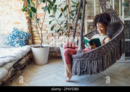 Young beautiful afro american girl is reading in a hanging wicker chair. Dark skinned teen in casual clothes is resting at home. Female teenage smilin Stock Photo