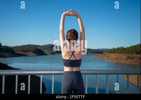 Rear view of a fit sporty woman doing and arm stretch in the sun by a lake Stock Photo
