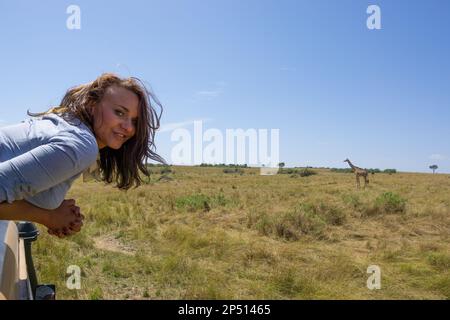Young female tourist looking out of a safari vehicle with a giraffe and beautiful landscape in the background  in masai mara kenya Stock Photo