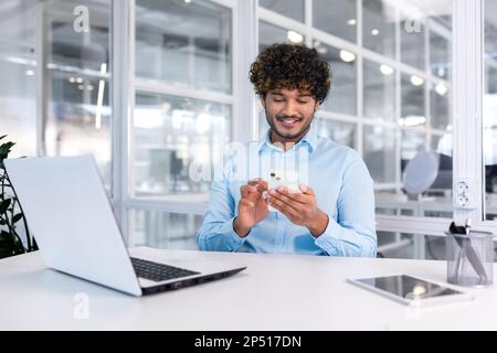Young indian programmer inside modern office sitting at workplace, man using phone, testing online application on new software, businessman entrepreneur in shirt smiling and happy with achievement. Stock Photo