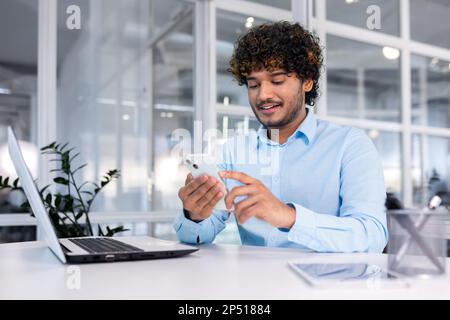 Young indian programmer inside modern office sitting at workplace, man using phone, testing online application on new software, businessman entrepreneur in shirt smiling and happy with achievement. Stock Photo