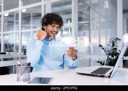 Young successful businessman inside the office at work uses a laptop hispanic man holds a smartphone in his hands, the man holds his hand up a gesture of success and triumph, celebrating victory. Stock Photo