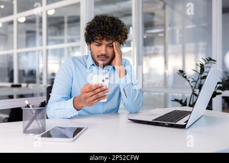 Businessman inside the office at the workplace is not satisfied with the work of the phone, the man is upset holding a smartphone in his hands, hispanic man is sitting at the table with a laptop. Stock Photo