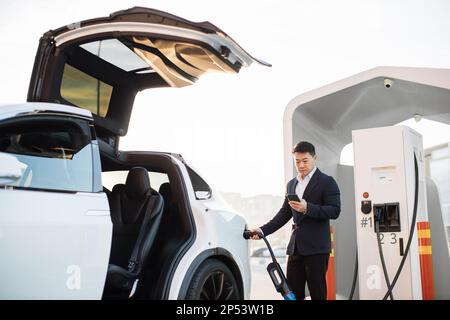 Asian businessman dressed in formal suit using smartphone app while charging luxury electric car on station. Confident man standing outdoors and plugging in power cord with cell phone in one hand. Stock Photo
