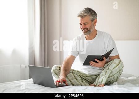 Cheerful mature man using laptop, taking notes in notepad Stock Photo