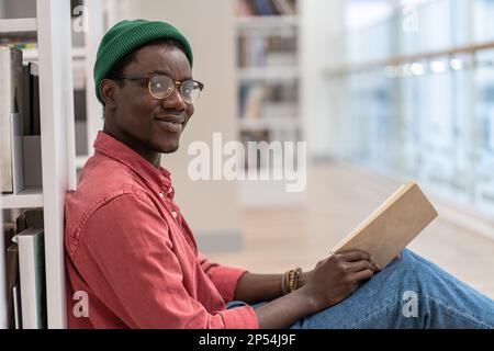 Black student man library visitor sitting on floor near bookcase with book in hands, looks at camera Stock Photo