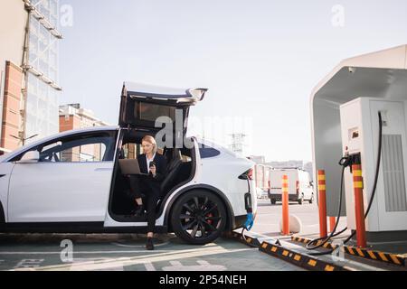 Caucasian business lady drinking takeaway coffee and working on wireless laptop while charging battery of her luxury electric car. Well dressed woman doing some business at public EV station. Stock Photo