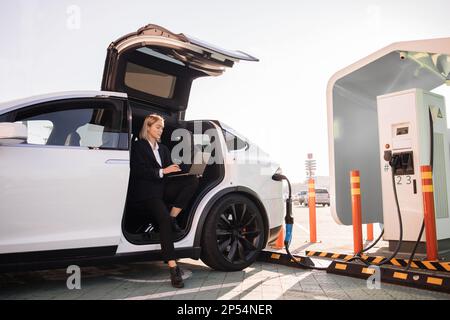 Caucasian business lady drinking takeaway coffee and working on wireless laptop while charging battery of her luxury electric car. Well dressed woman doing some business at public EV station. Stock Photo